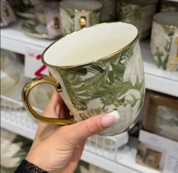 Porcelanowy kubek z motywem zielonych liści i kwiatów, ozdobiony złotym rantem i uchwytem, trzymany w dłoni przed regałem z innymi naczyniami o podobnym wzorze.