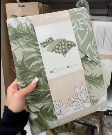 Pościel z motywem zielonych liści i białych kwiatów, trzymana w dłoni, z widocznym opakowaniem prezentującym grafikę wzoru oraz informacje o wymiarach kompletu.