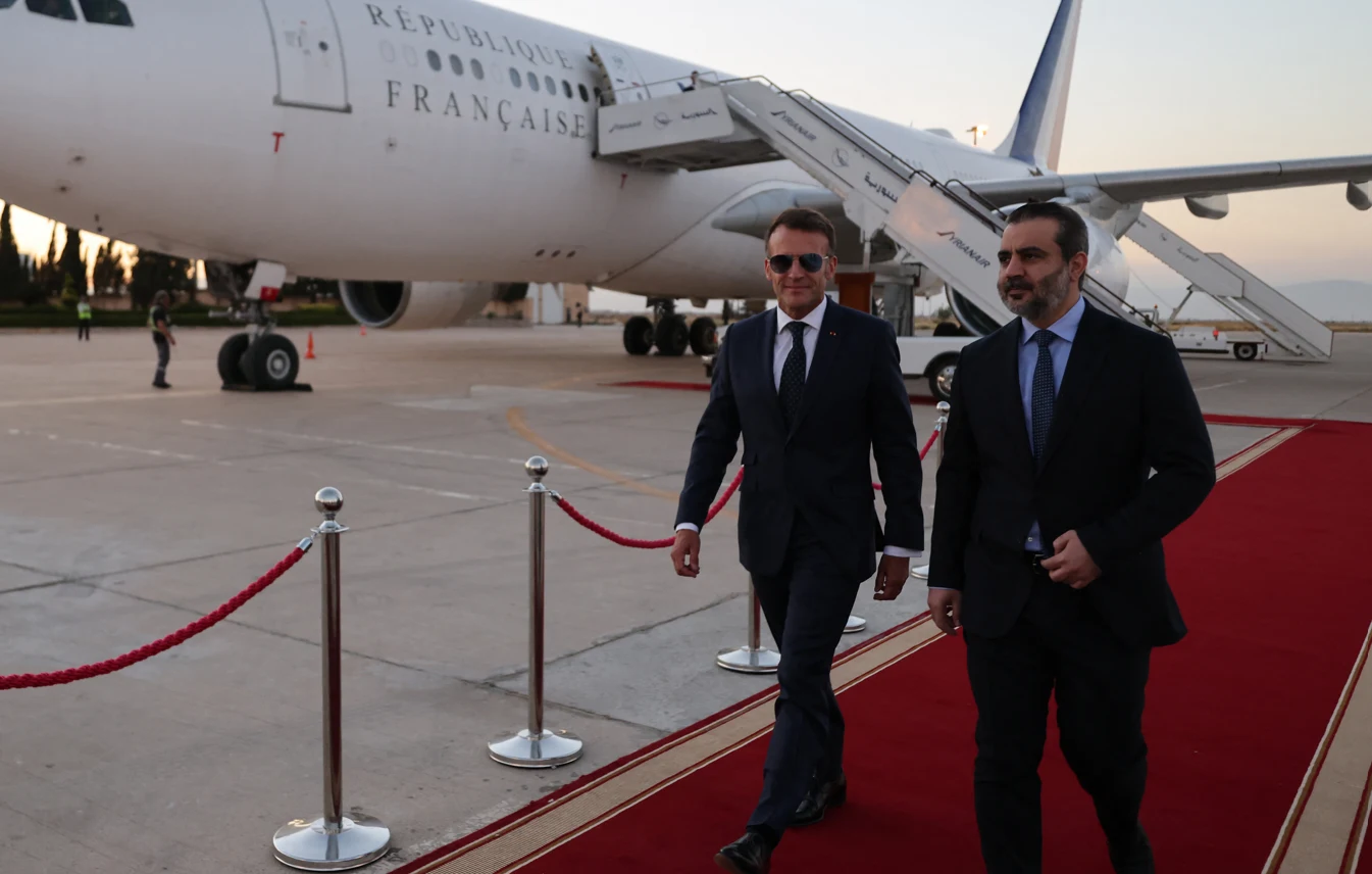 Dwóch elegancko ubranych mężczyzn idzie po czerwonym dywanie na tle samolotu z napisem 'République Française', prowadząc oficjalną delegację na lotnisku.