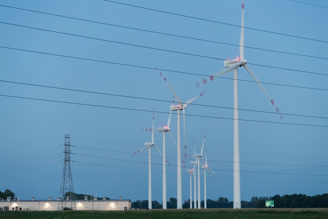 Rada Ministrów przyjęła projekt nowelizacji ustawy o odnawialnych źródłach energii Rząd turbin wiatrowych na tle nieba, przy budynku przemysłowym i liniach energetycznych.