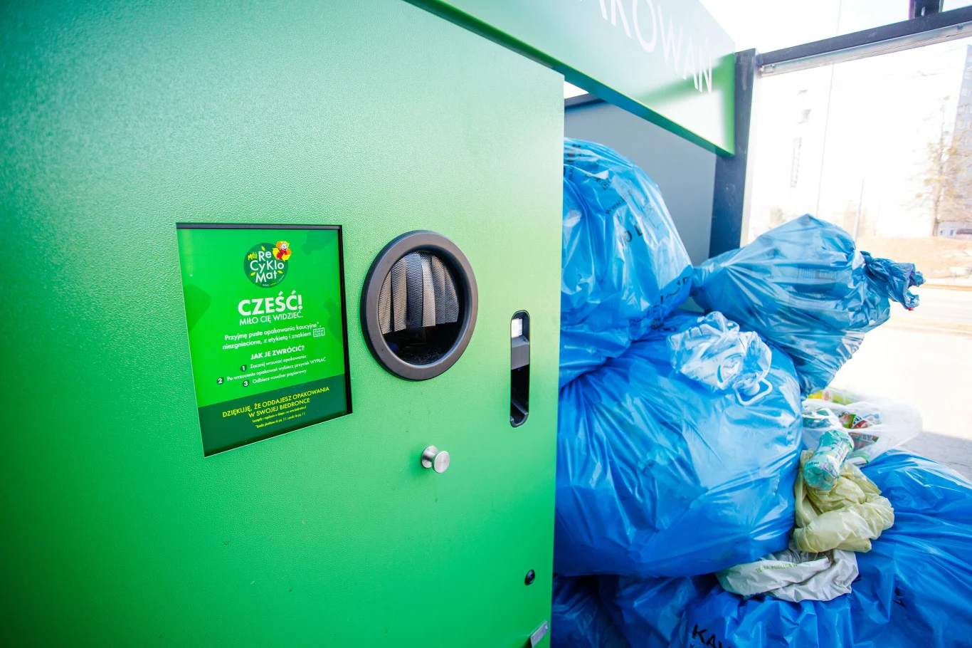 Automat do przyjmowania butelek i puszek obok dużej ilości niebieskich worków wypełnionych odpadami, ustawionych przy wejściu do punktu recyklingu.