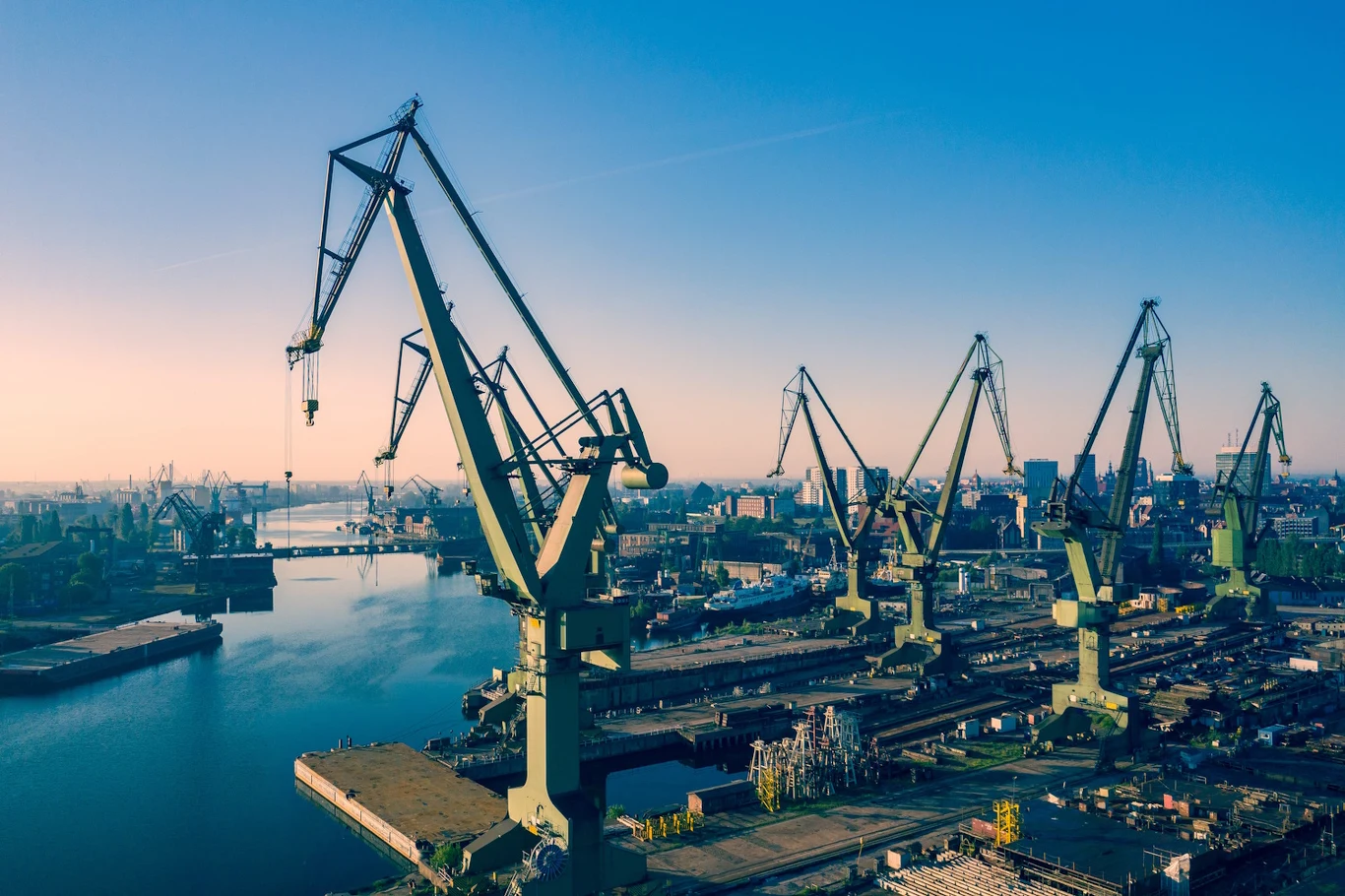 Rząd wysokich dźwigów portowych rozciągających się wzdłuż nabrzeża stoczniowego z widokiem na spokojną taflę wody i panoramę zabudowań przemysłowych w tle.
