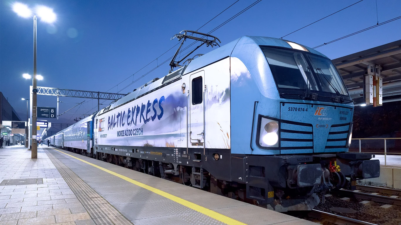 Pociąg pasażerski Baltic Express z nowoczesną, niebieską lokomotywą stoi przy peronie oświetlonej stacji kolejowej.