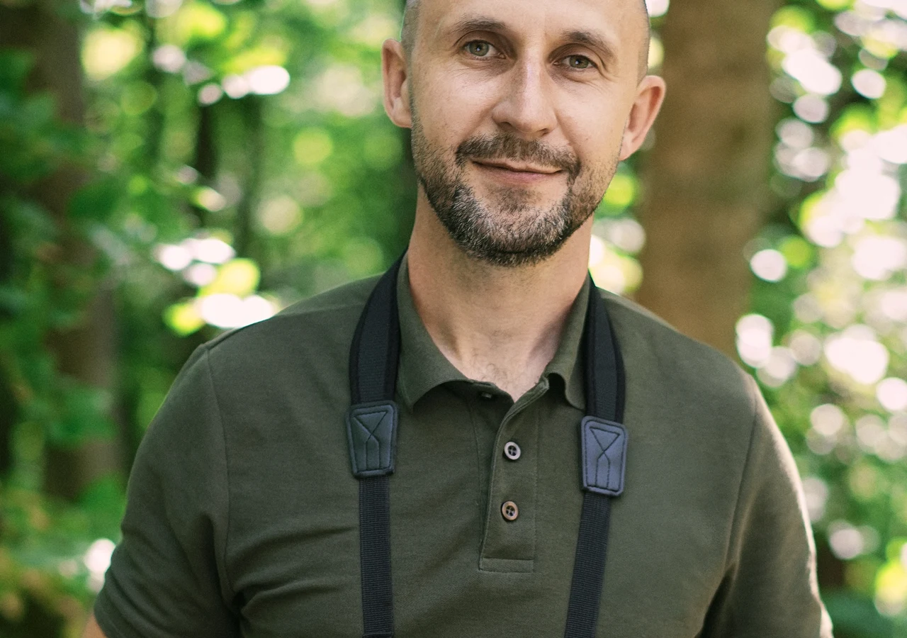 Maciej, kandydat z programu "Wielka Miłość w Małym Mieście" Uśmiechnięty mężczyzna w zielonej koszulce polo z czarnymi paskami aparatu na szyi stoi w lesie, otoczony rozmytym tłem drzew i zieleni.