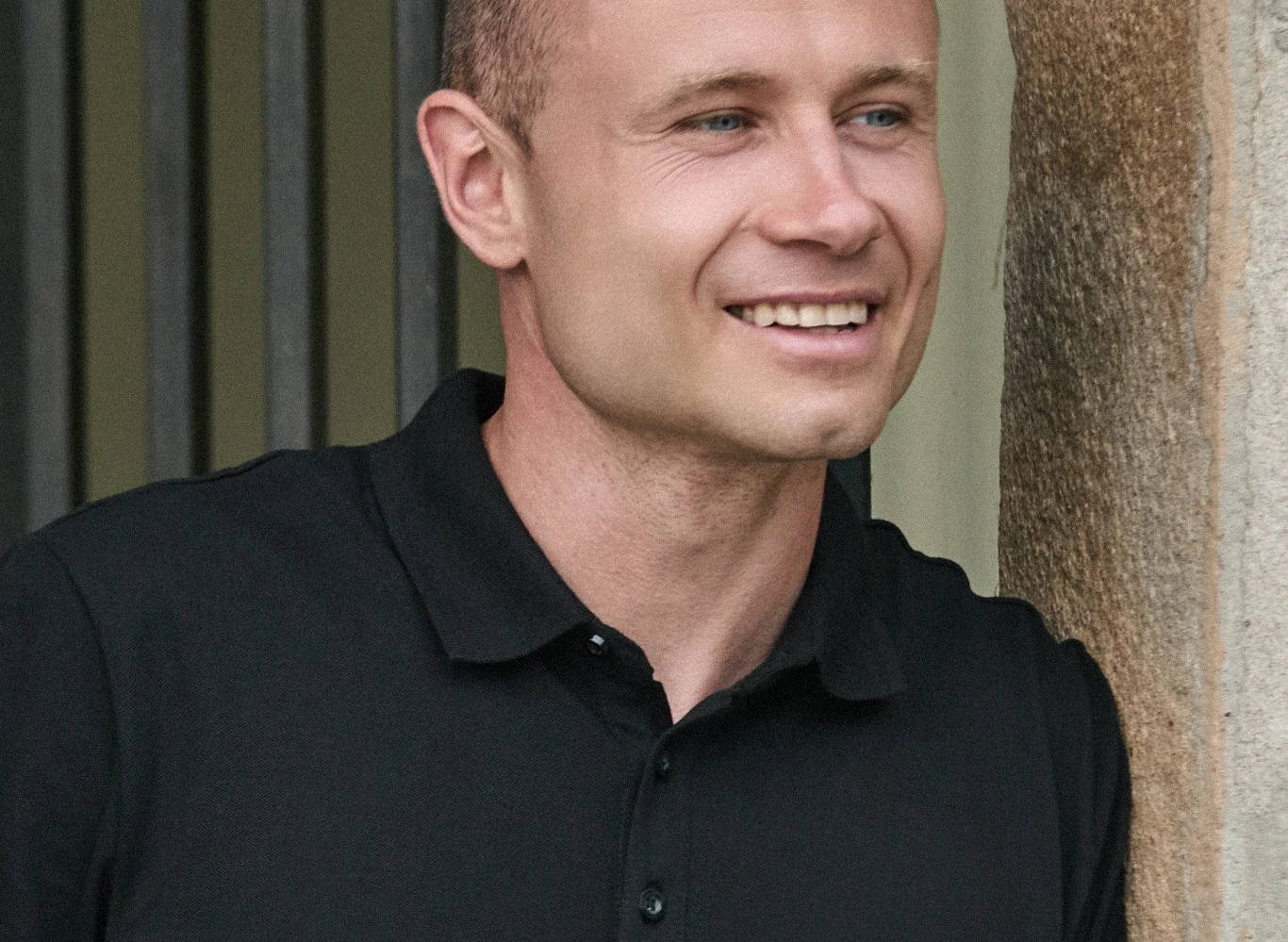 Rafał, kandydat z programu "Wielka Miłość w Małym Mieście" Mężczyzna w czarnej koszulce polo oparty o ścianę, uśmiechnięty, z krótkimi włosami.