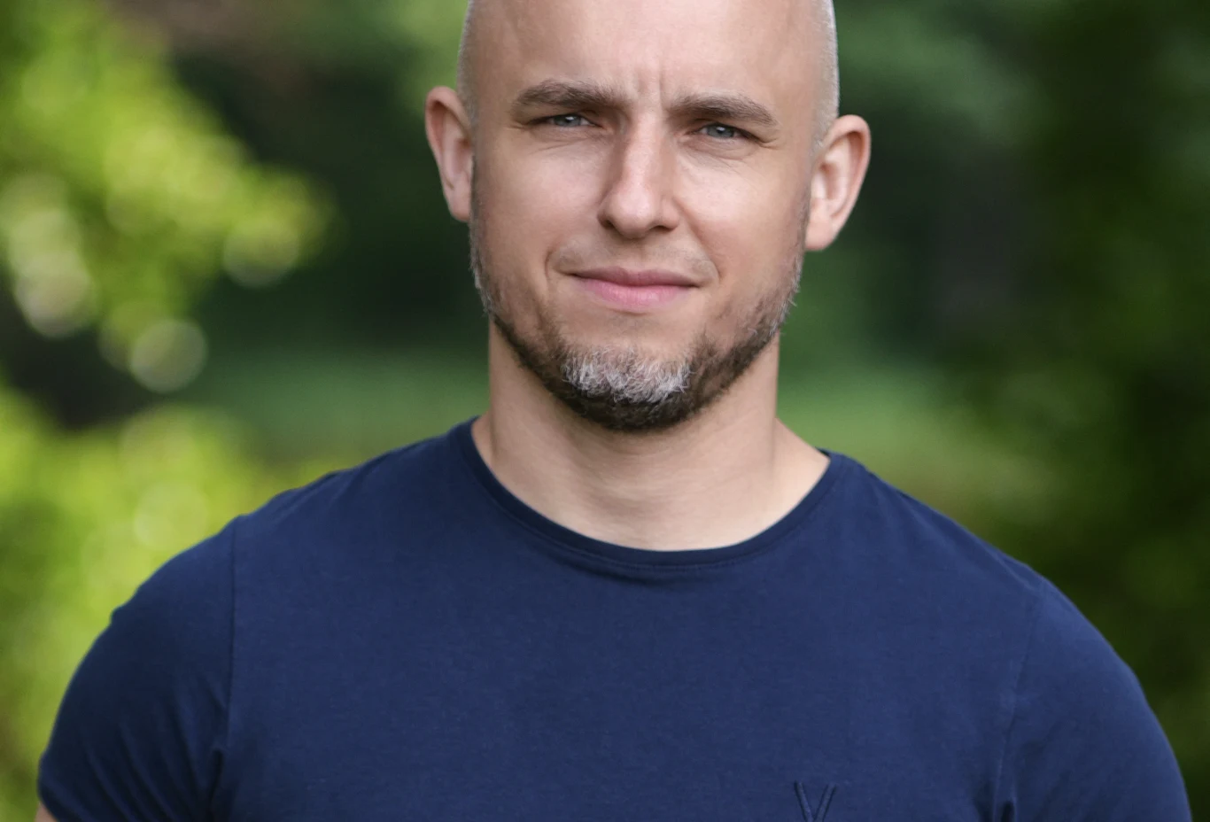 Jarek, kandydat z programu "Wielka Miłość w Małym Mieście" Uśmiechnięty mężczyzna z krótkimi, jasnymi włosami i zarostem, ubrany w granatowy t-shirt, stoi na tle zieleni.