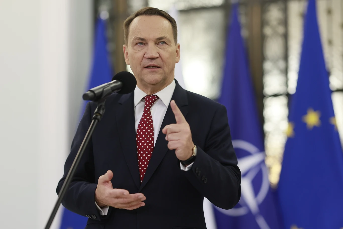 Szef MSZ Radosław SIkorski Radosław Sikorski w garniturze i czerwonej krawacie mówi do mikrofonu na tle flag NATO i Unii Europejskiej.