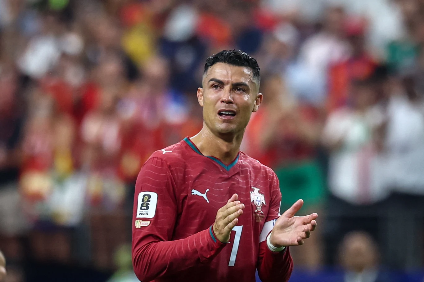Cristiano Ronaldo Piłkarz w czerwonej koszulce z widocznym numerem 7 na piersi, z wyrazem silnych emocji na twarzy, klaszczący na tle pełnych trybun stadionu.