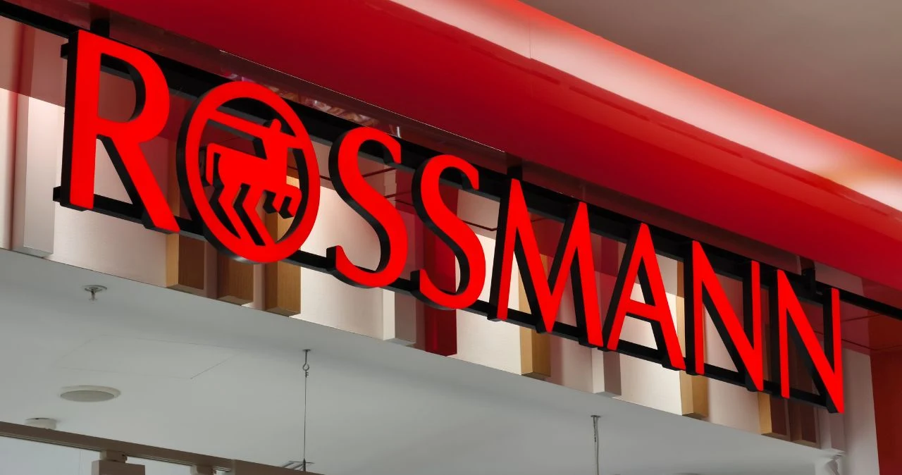 Rossmann mocno przecenił kosmetyki do włosów. Litr szamponu kupisz za 15,49 zł Logo Rossmann w czerwieni z symbolem centaura na tle białej i czerwonej ściany, widoczne oznakowanie sklepu.
