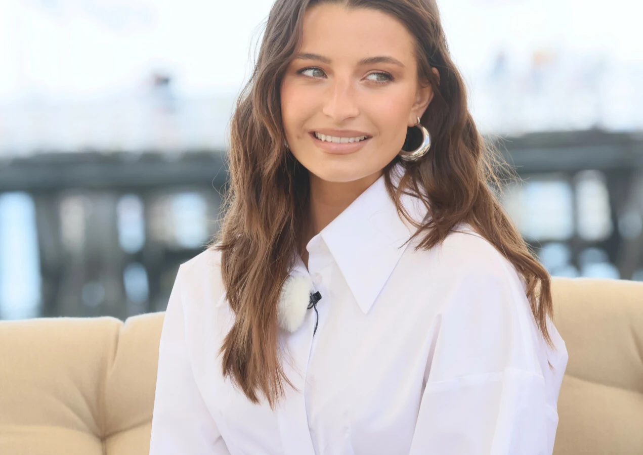 Julia Wieniawa Uśmiechnięta kobieta z długimi, brązowymi włosami, ubrana w białą koszulę z dużym kołnierzem i srebrne kolczyki, siedząca na jasnej sofie.
