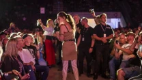 Kobieta z mikrofonem w centrum uwagi, otoczona przez ochroniarzy i publiczność podczas wydarzenia na dużej sali, widzowie fotografują i nagrywają scenę telefonami.