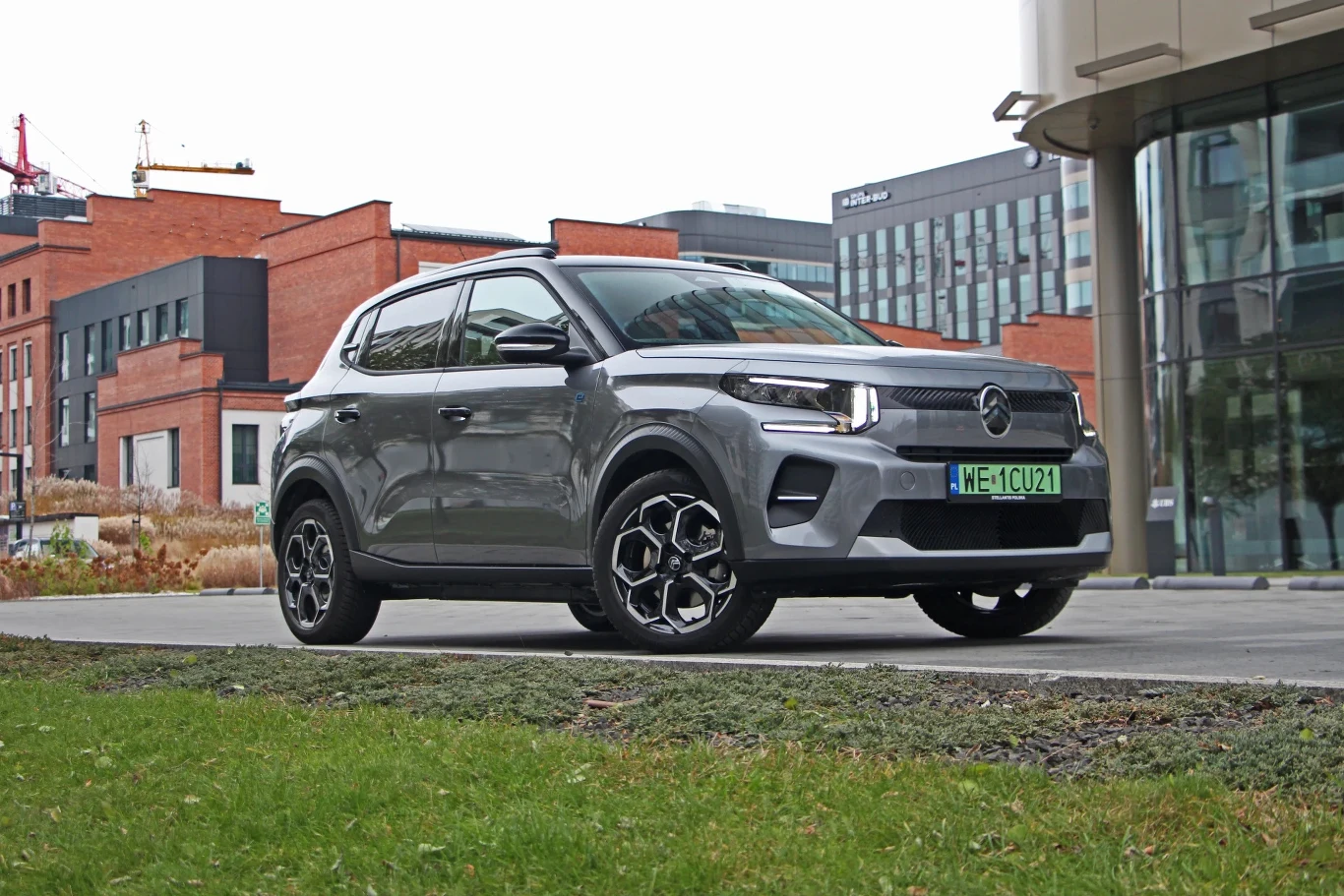 Srebrny samochód typu SUV z nowoczesnym designem i charakterystycznymi czarnymi felgami stoi zaparkowany na tle miejskich budynków z czerwonej i szklanej cegły.