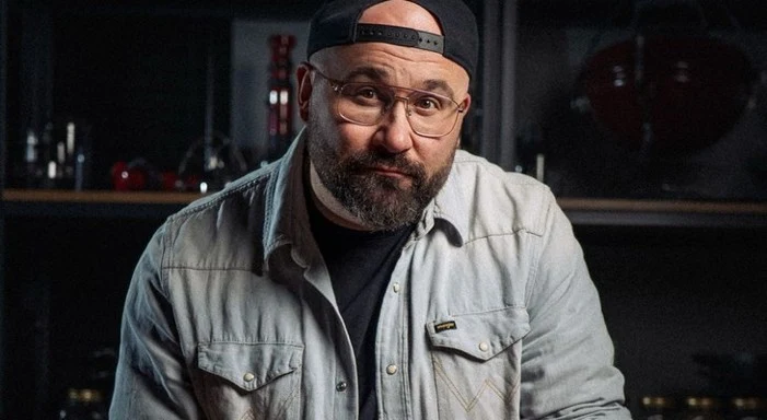 Brodaty mężczyzna w jasnej koszuli i czapce z daszkiem założonej tyłem, patrzący poważnie w stronę obiektywu.