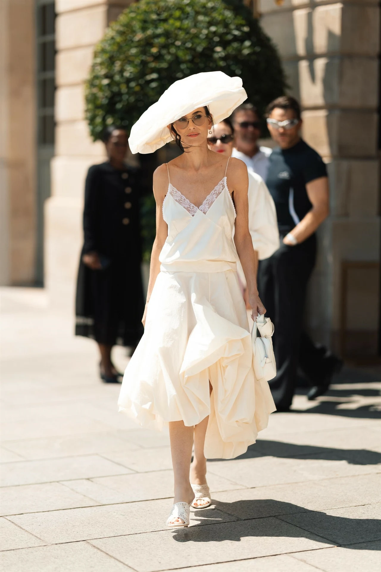 Demi Moore w stylizacji Magdy Butrym. Demi Moore w eleganckiej, jasnej sukience midi i dużym, szerokim kapeluszu, niosąca jasną torebkę, czyli stylizacji przygotowanej przez Magdę Butrym, idzie chodnikiem w słoneczny dzień.