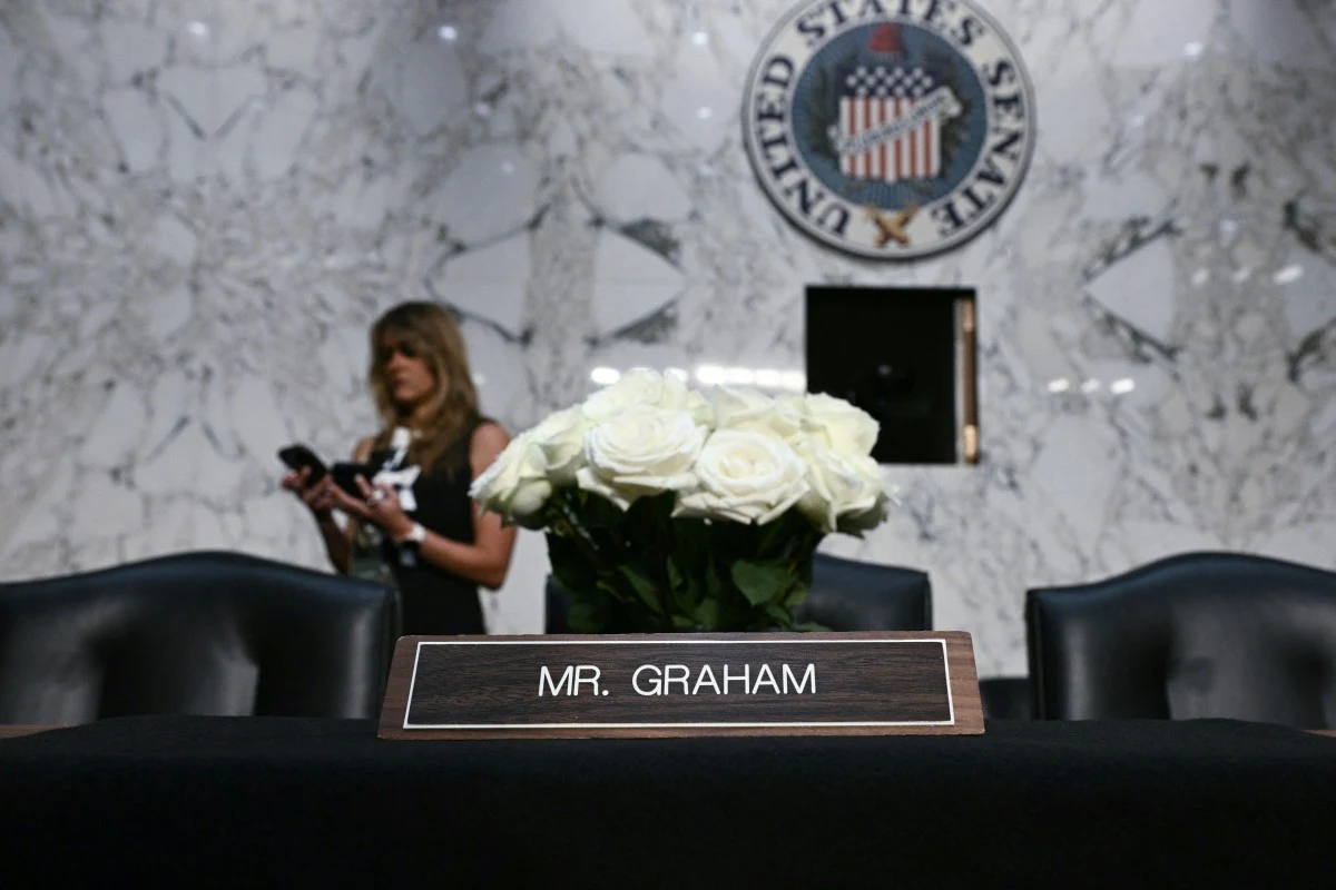 Tabliczka z napisem „Mr. Graham” ustawiona na czarnym stole, na którym znajduje się również bukiet białych róż, w tle marmurowa ściana i herb Senatu Stanów Zjednoczonych.