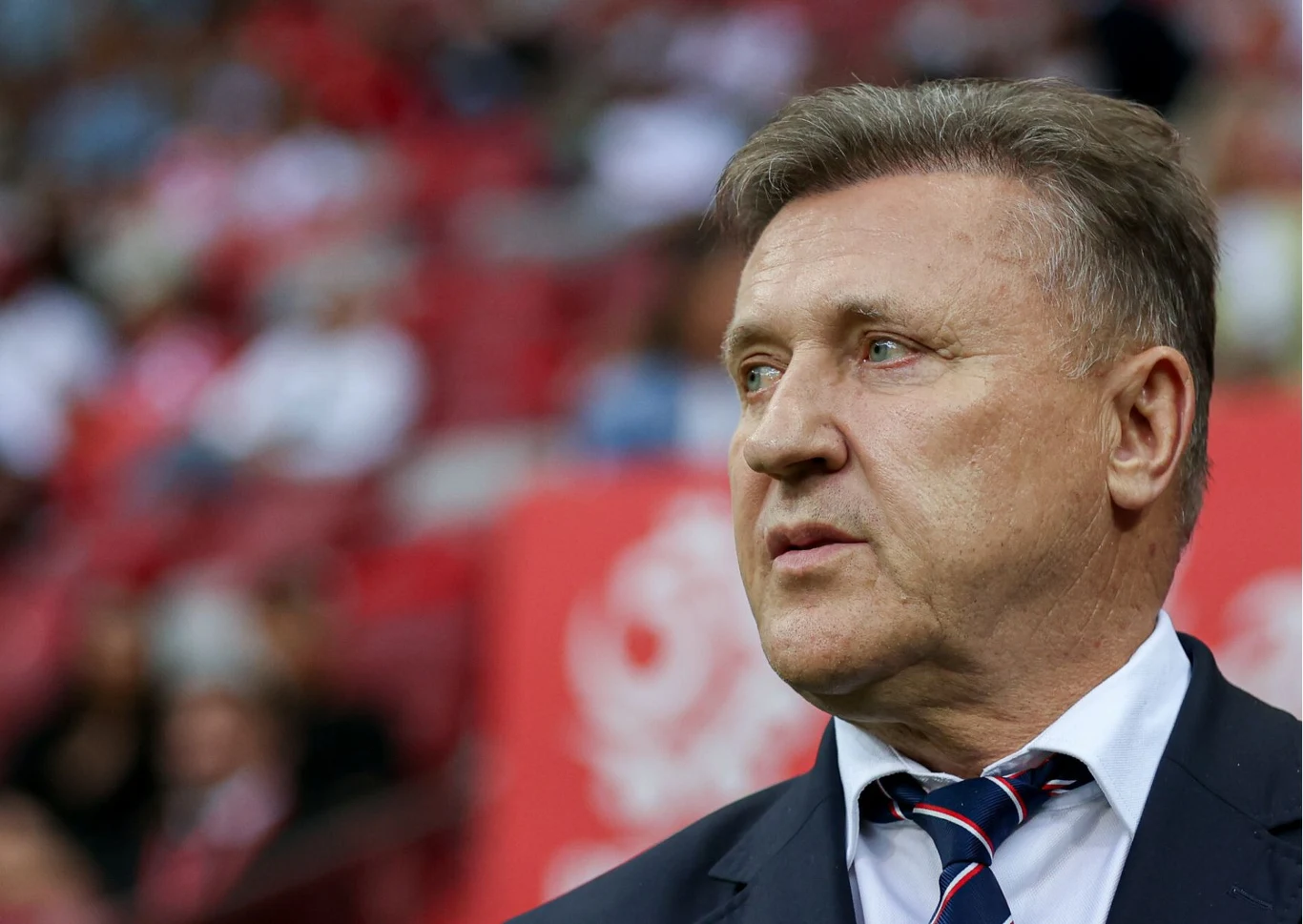 Mężczyzna w garniturze i krawacie patrzący w bok, na tle rozmytego czerwonego stadionu z wyraźnym symbolem białego orła.