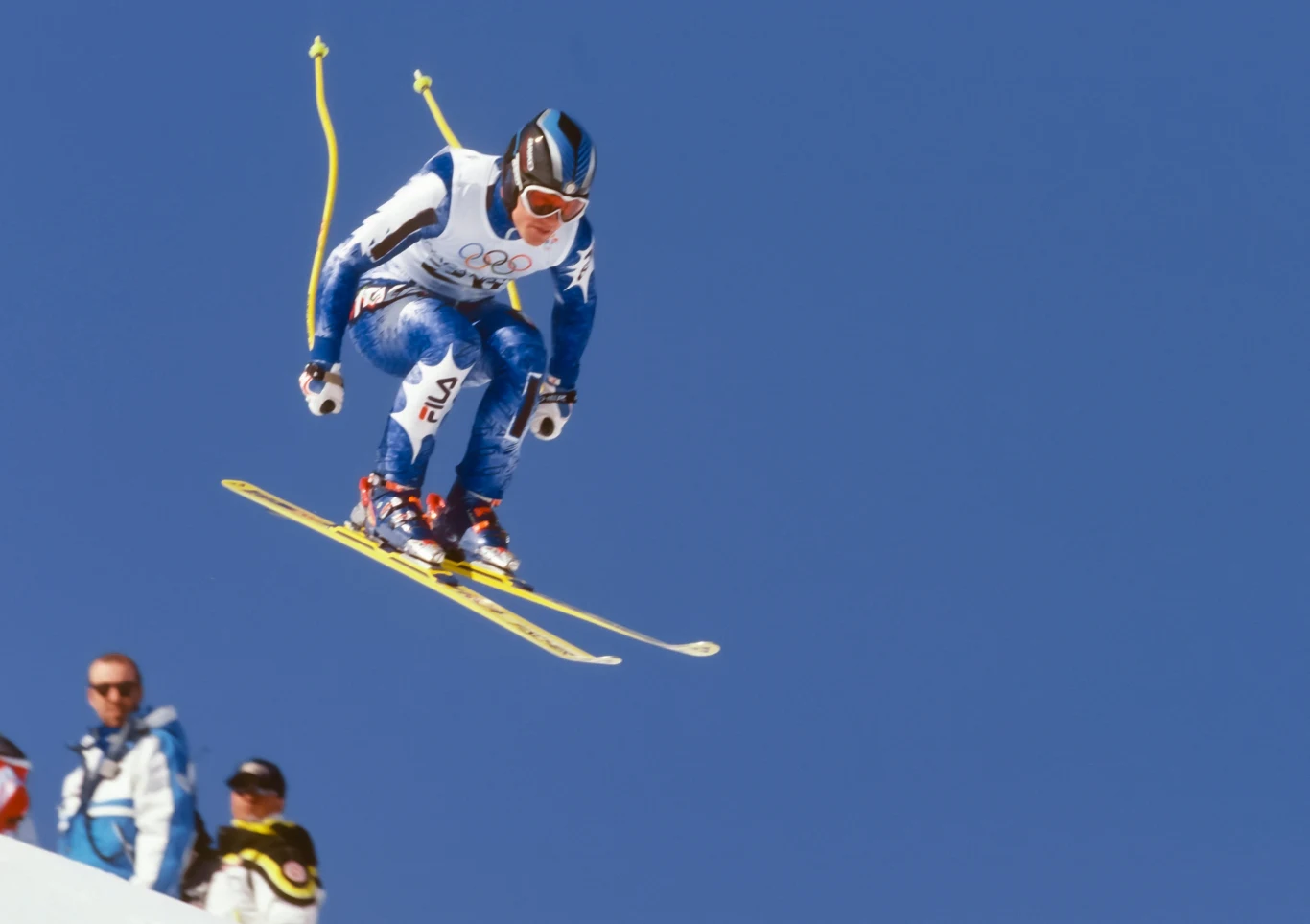 Narciarz w kombinezonie olimpijskim wykonuje skok na nartach zjazdowych na tle bezchmurnego, niebieskiego nieba.