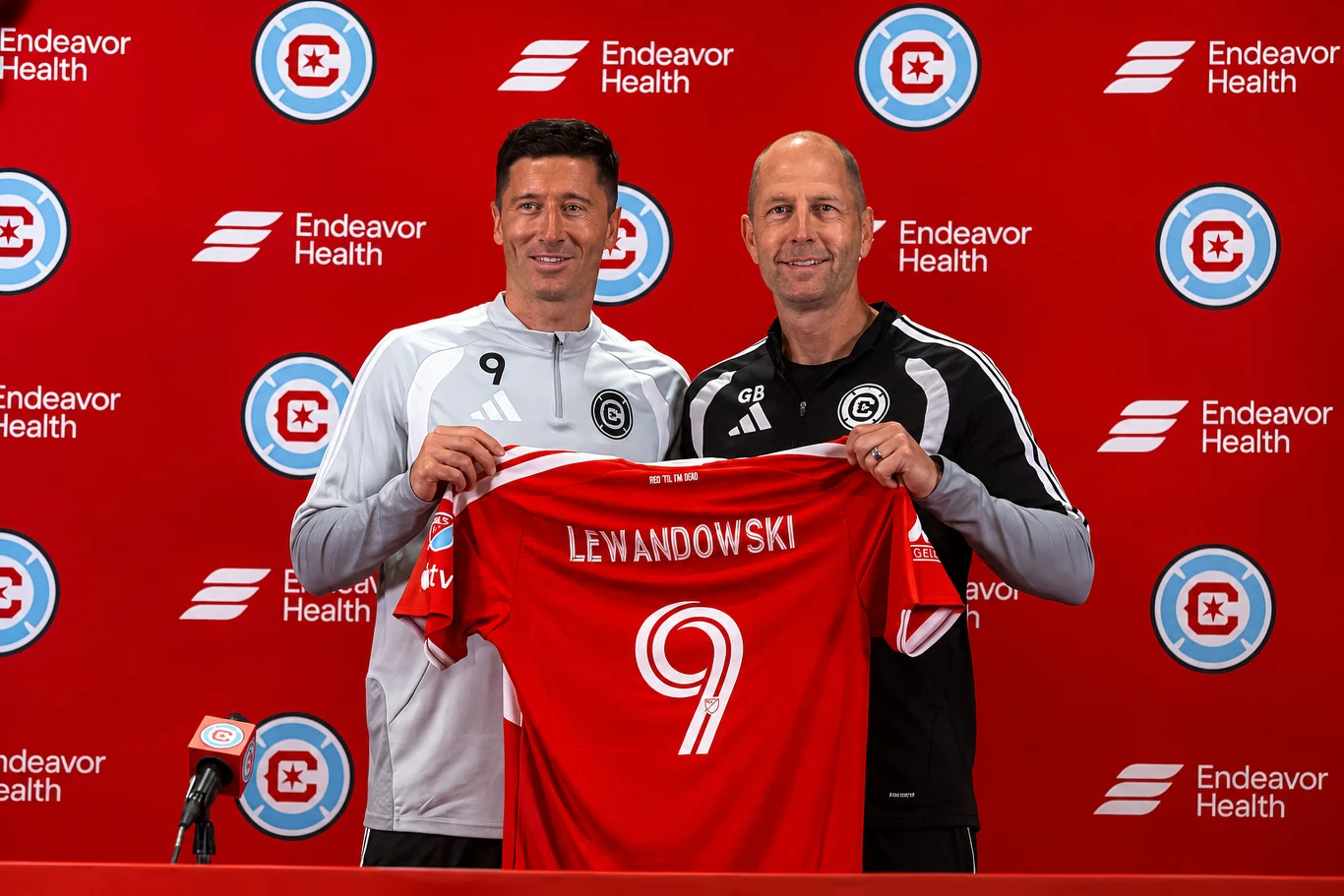 Dwóch mężczyzn trzymających czerwoną koszulkę piłkarską z nazwiskiem Lewandowski i numerem 9 na tle ściany z logo klubu sportowego i sponsora.