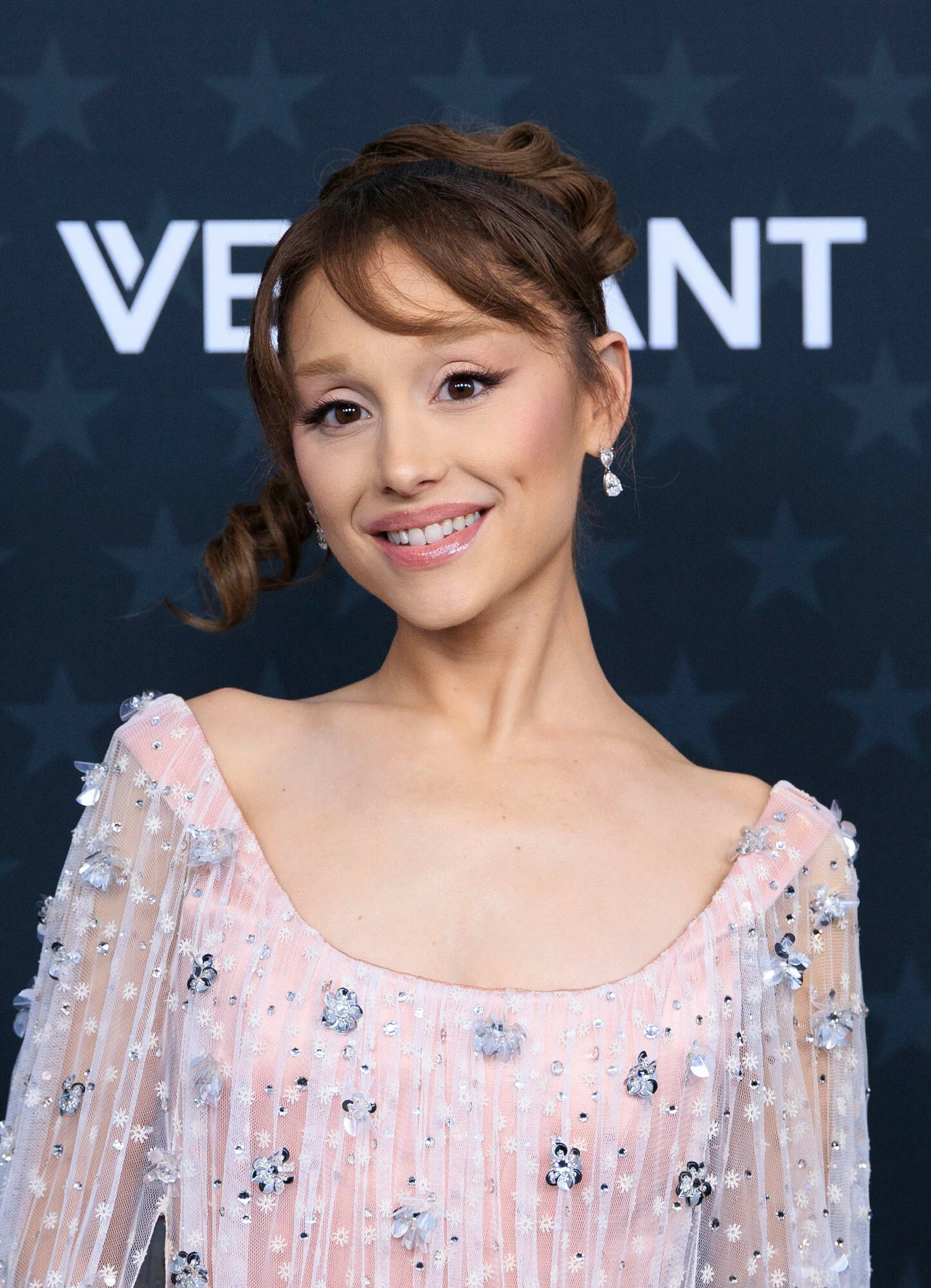 Ariana Grande Ariana Grande z upiętymi włosami w eleganckiej, jasnoróżowej sukni ozdobionej kwiatowymi aplikacjami, stojąca na tle ze wzorem gwiazd.