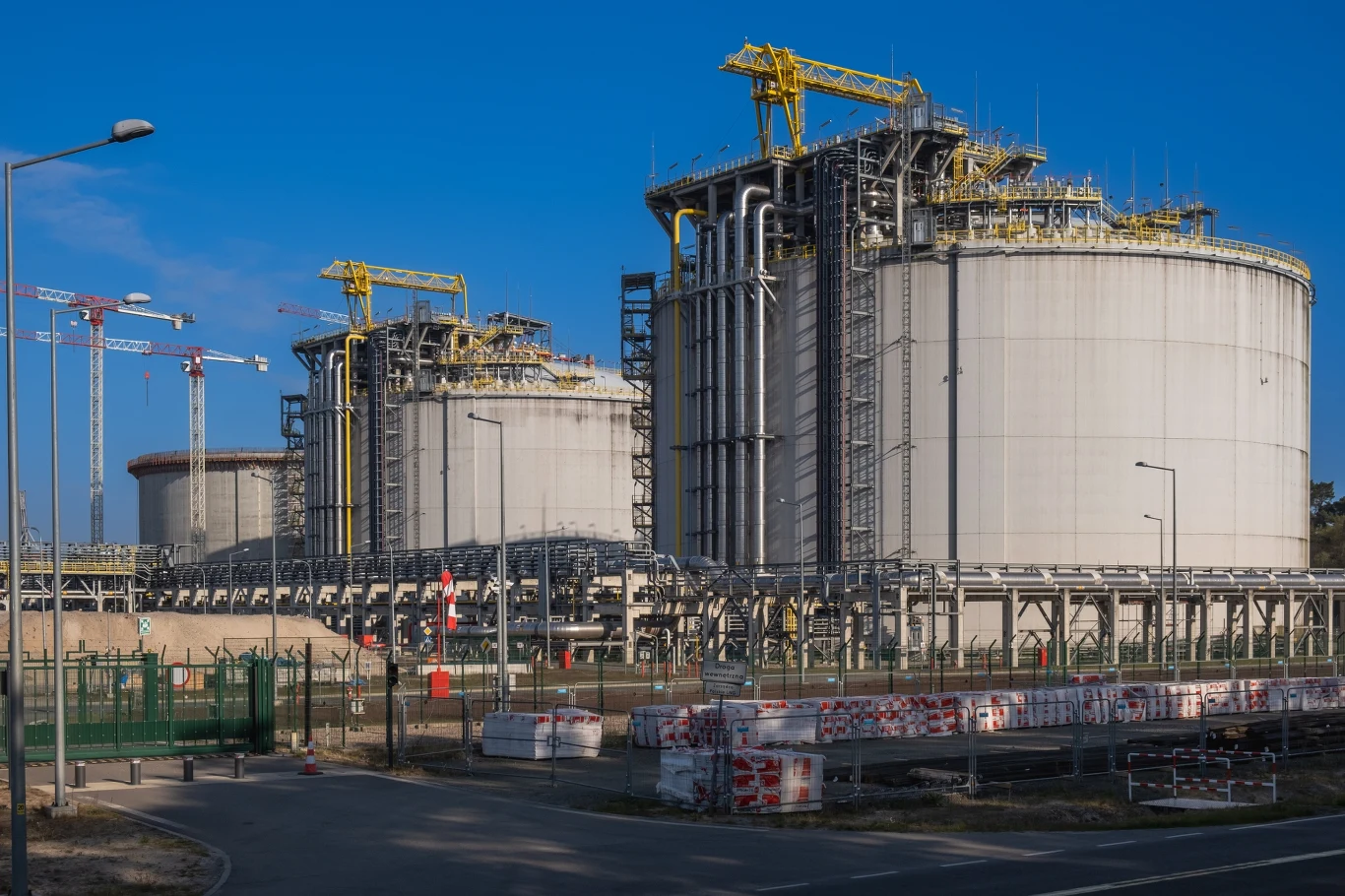 Dwie ogromne cylindryczne zbiorniki przemysłowe otoczone infrastrukturą techniczną, żółtymi suwnicami oraz ogrodzeniem, w tle kilka wysokich dźwigów budowlanych na niebieskim niebie.