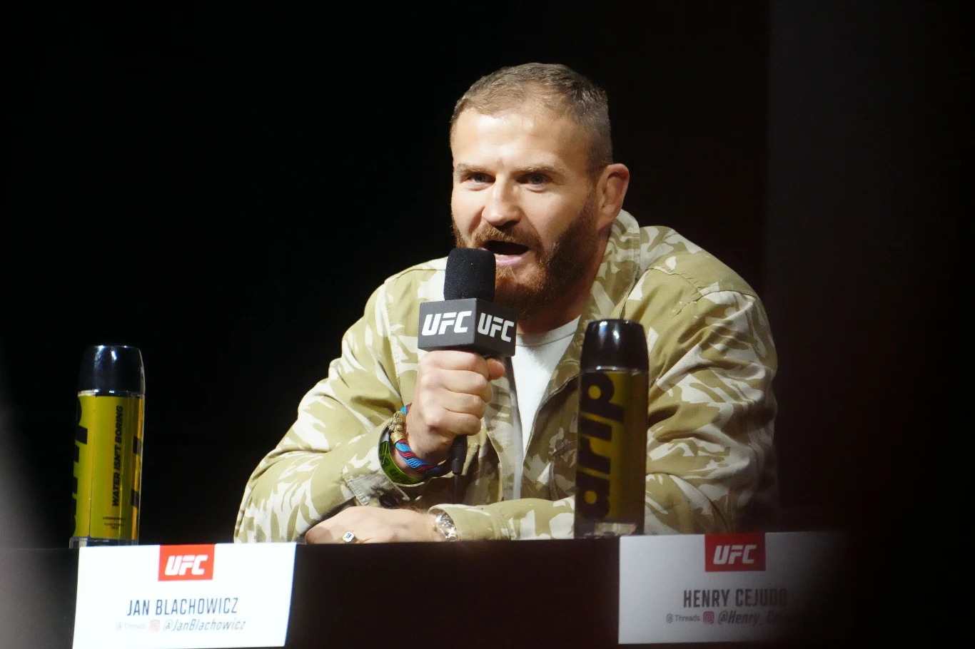 Mężczyzna w jasnej, wzorzystej kurtce siedzi za stołem z mikrofonem UFC w ręku, rozmawia podczas konferencji prasowej, po bokach widoczne są butelki z napojem i tabliczka z imieniem oraz nazwiskiem.