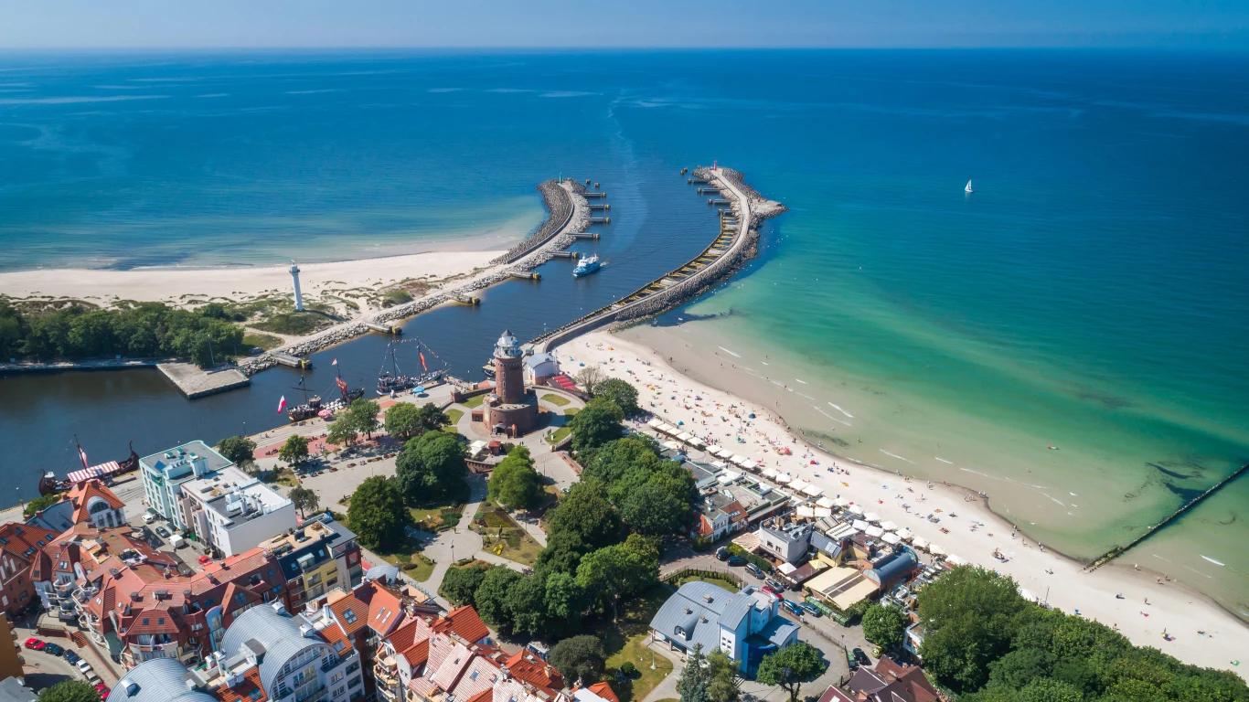 Latarnia morska otoczona zielenią, szeroka plaża z licznymi plażowiczami oraz dwa falochrony wysunięte w morze rozdzielające ujście rzeki do Bałtyku.