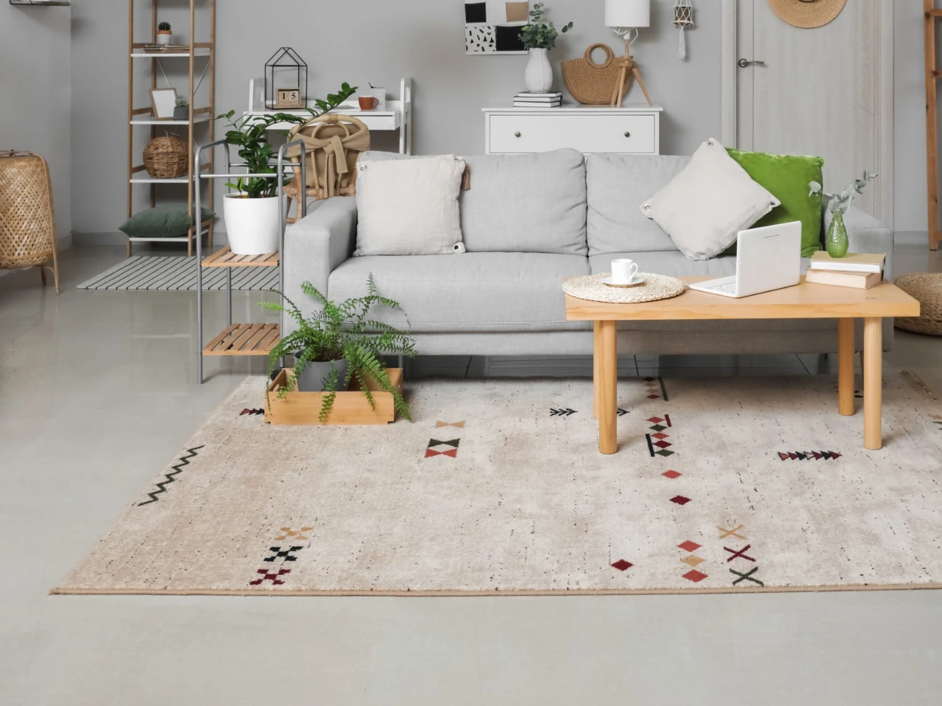Jasnoszara sofa z poduszkami, drewniany stolik z laptopem oraz filiżanką, dywan z geometrycznym wzorem, drewniana skrzynka z paprocią i minimalistyczne dekoracje w tle.