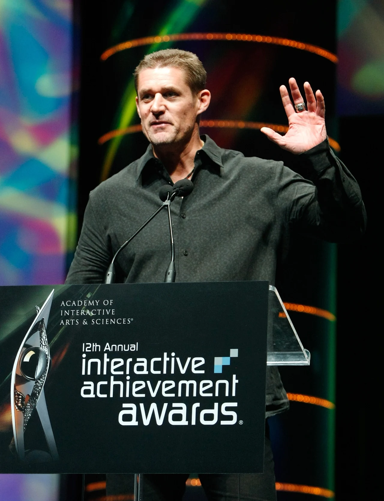 Mężczyzna w ciemnej koszuli stoi za mównicą z logo 'interactive achievement awards', mówi do mikrofonu i unosi prawą rękę w geście powitania, w tle wielokolorowe światła i dekoracje sceniczne.