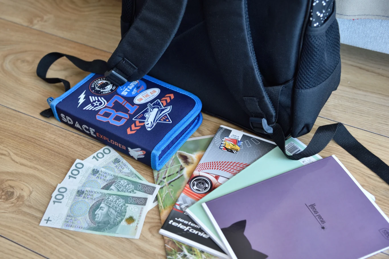 Stypendium dla ucznia na rok szkolny 2026/2027 Czarny plecak, niebieski piórnik z motywem kosmosu, banknoty 100 zł, zeszyty i podręczniki na podłodze.