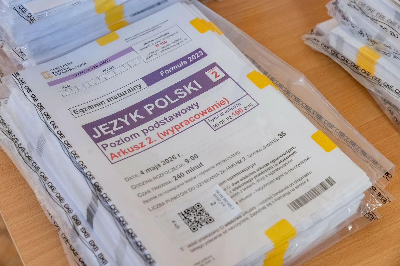 Stos egzemplarzy arkuszy maturalnych egzaminu z języka polskiego na poziomie podstawowym, zapakowanych w przezroczyste foliowe opakowanie i oznaczonych żółtymi zabezpieczeniami.
