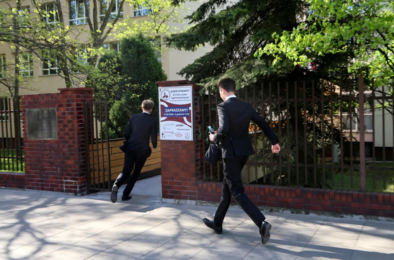 Dwóch młodych mężczyzn w garniturach biegnie w stronę wejścia do budynku otoczonego ogrodzeniem z czerwonej cegły, w tle widoczne są drzewa i plakat informacyjny przy bramie.