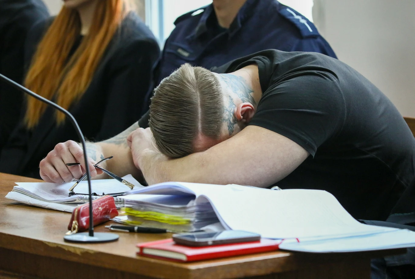 Mężczyzna z tatuażem na szyi siedzi przy stole w sali sądowej, opierając głowę na rękach obok rozłożonych dokumentów i okularów.