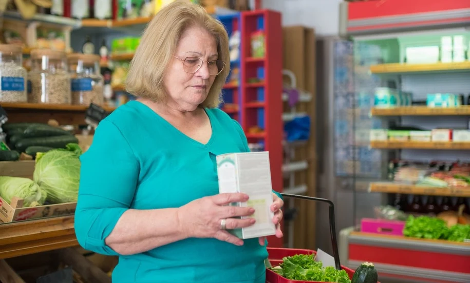 Zakupy po 60-tce — zniżki z kartą seniora Starsza kobieta z koszykiem czyta etykietę produktu w sklepie spożywczym — zakupy z kartą seniora