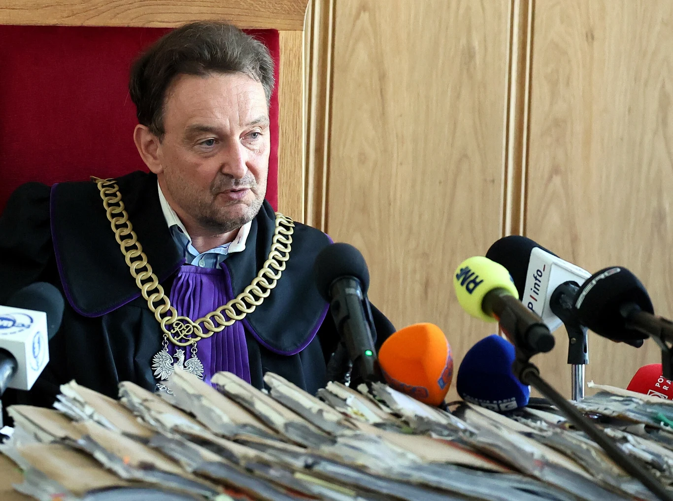 Sędzia w todze i łańcuchu z orłem, siedzący przy stole z licznymi teczkami akt oraz mikrofonami ustawionymi przed nim.