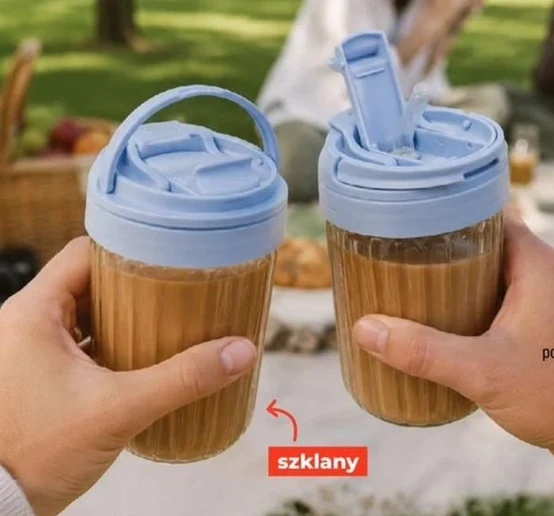 Dwa szklane kubki z mleczną kawą i niebieskimi pokrywkami, trzymane w dłoniach, na tle pikniku z koszem i rozłożonym kocem.