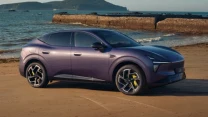 Fioletowy nowoczesny samochód elektryczny typu SUV zaparkowany na piaszczystej plaży z widokiem na spokojne morze i zielone wzgórza w oddali.