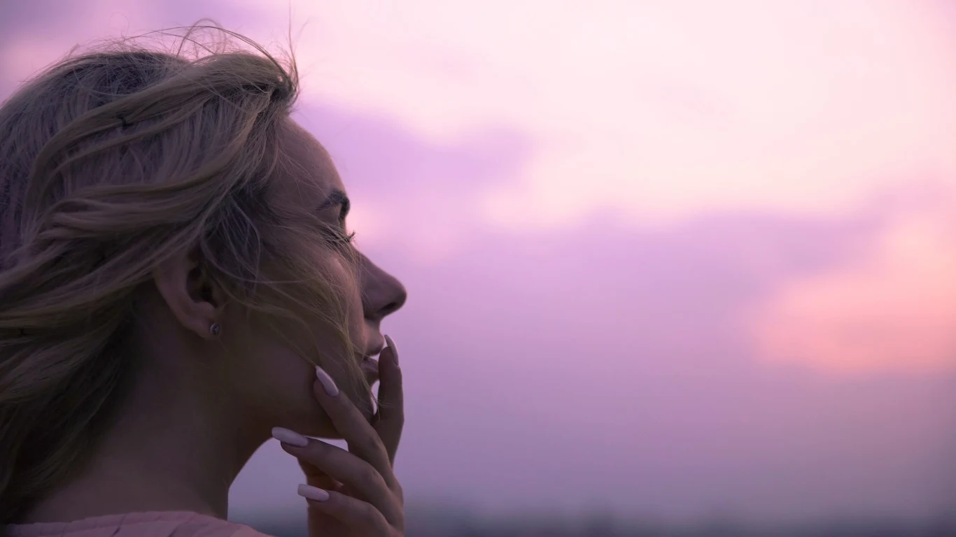 Astrologia od dawna przygląda się wpływowi Księżyca na nasze życie Kobieta z blond włosami patrzy zamyślona w stronę różowo-fioletowego nieba o zachodzie słońca, delikatnie opierając dłoń na twarzy.