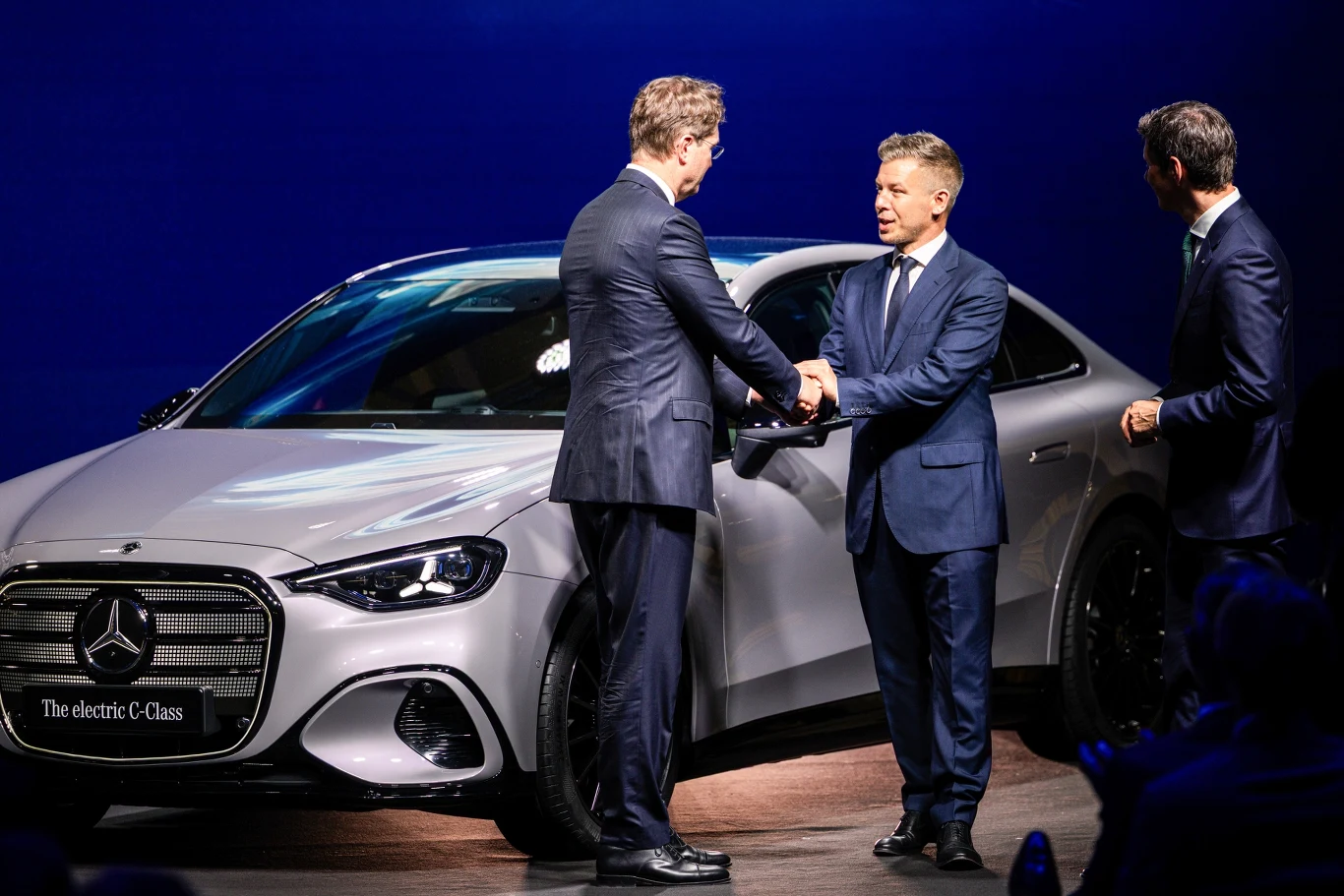 Trzech elegancko ubranych mężczyzn w garniturach wymienia uścisk dłoni i rozmowę przed nowoczesnym, srebrnym samochodem elektrycznym marki Mercedes na tle jasnego oświetlenia.