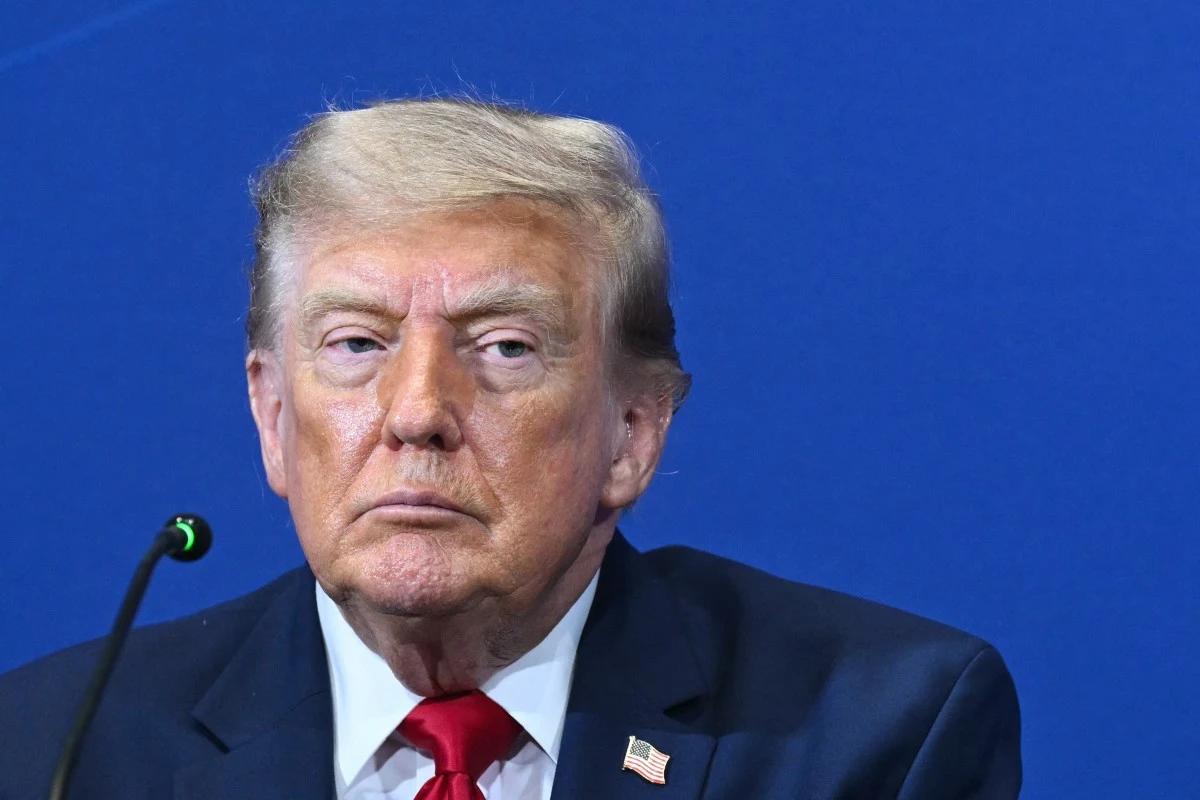 Donald Trump Mężczyzna w garniturze z amerykańską flagą w formie przypinki na klapie, siedzący przy mikrofonie na tle niebieskiej ściany.