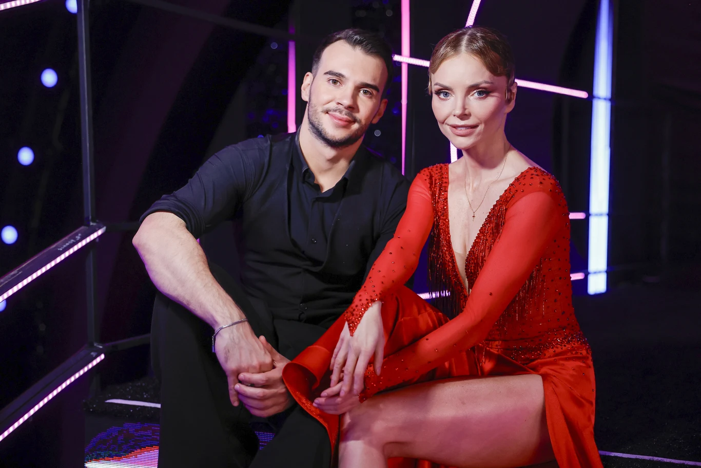 Albert Kosiński i Iza Miko w 18. edycji "Tańca z gwiazdami" Para elegancko ubranych osób siedzących obok siebie, mężczyzna w czarnej koszuli oraz kobieta w czerwonej, błyszczącej sukience z rozcięciem, otoczona kolorowymi światłami sceny.
