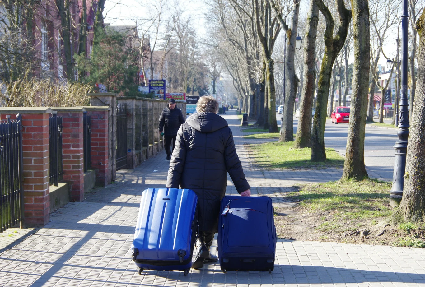 Ile się czeka na wyjazd do sanatorium w roku 2026? Sprawdź, jak sytuacja wygląda w poszczególnych województwach. Starsza kobieta w ciemnej kurtce, ciągnąca dwie duże walizki na chodniku wzdłuż ulicy. Wyjazd do sanatorium.