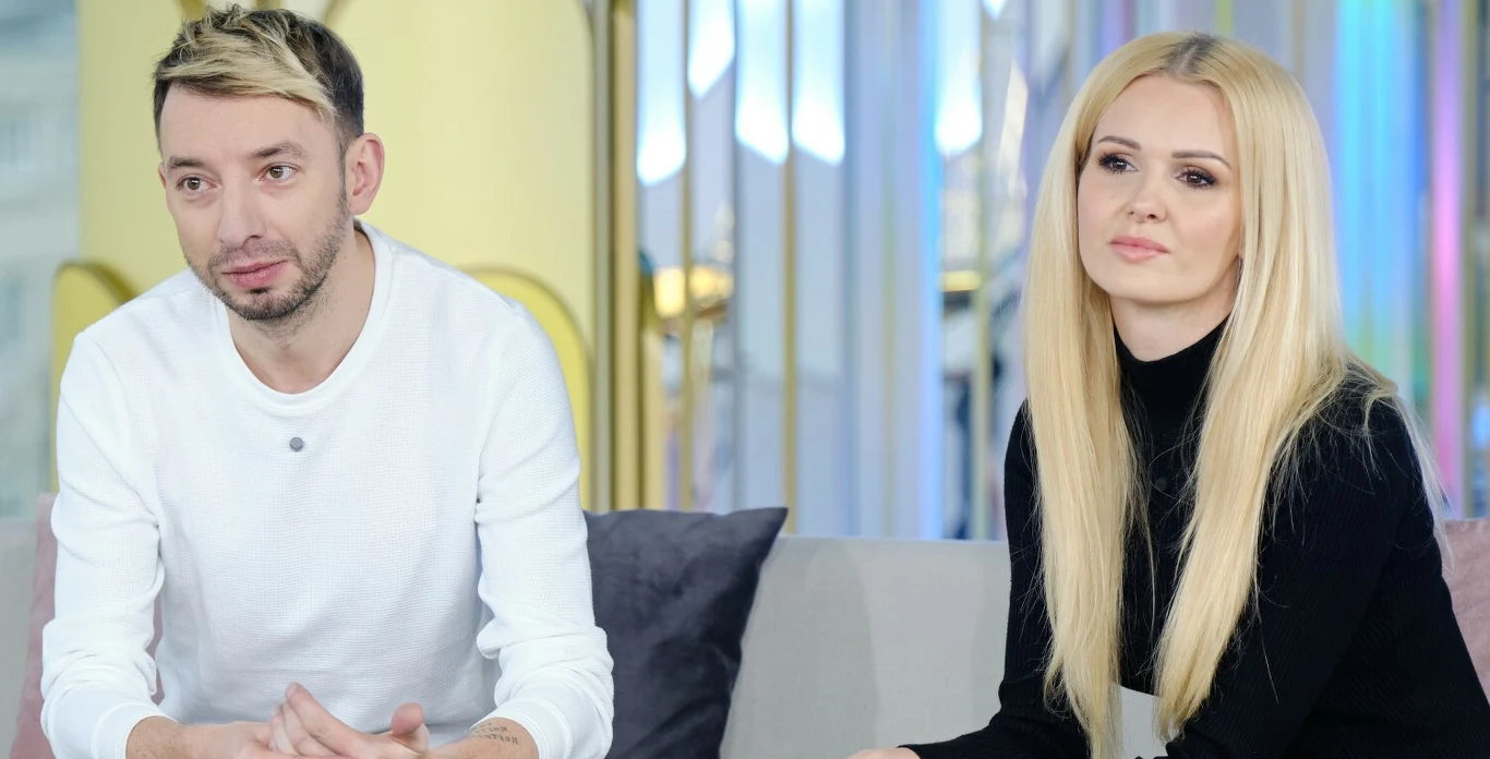 Adam Konkol i Angelina Konkol Dwoje dorosłych siedzących na kanapie, mężczyzna w białej koszulce z jasnymi włosami i kobieta o długich blond włosach w czarnym ubraniu, patrzący przed siebie w spokojnej atmosferze.