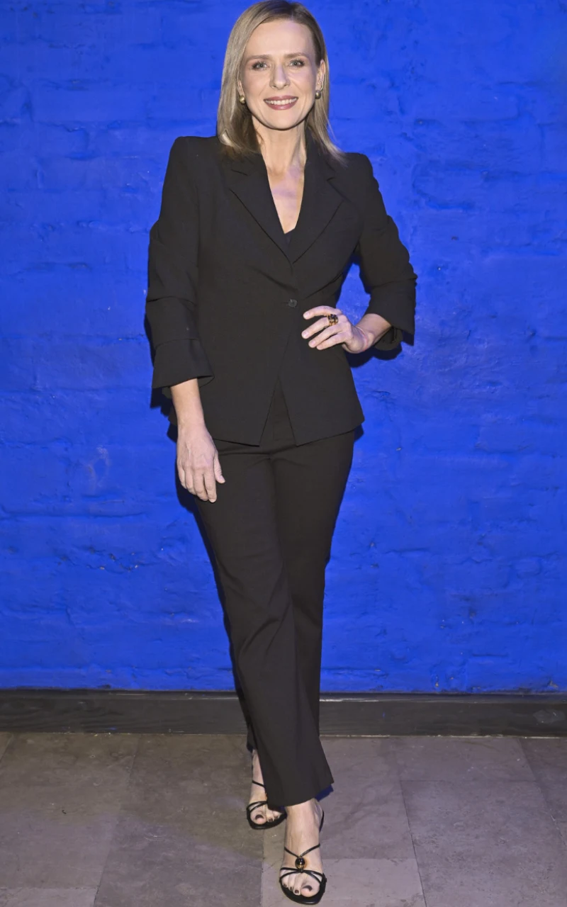Aneta Zając uwielbia garnitury. Tu, oficjalny i bardzo elegancki zestaw połączyła z sandałami na obcasie. Dzięki temu dodała stylizacji lekkości Kobieta w eleganckim, czarnym garniturze i sandałach na obcasie pozuje na tle intensywnie niebieskiej ściany, uśmiechając się do obiektywu.