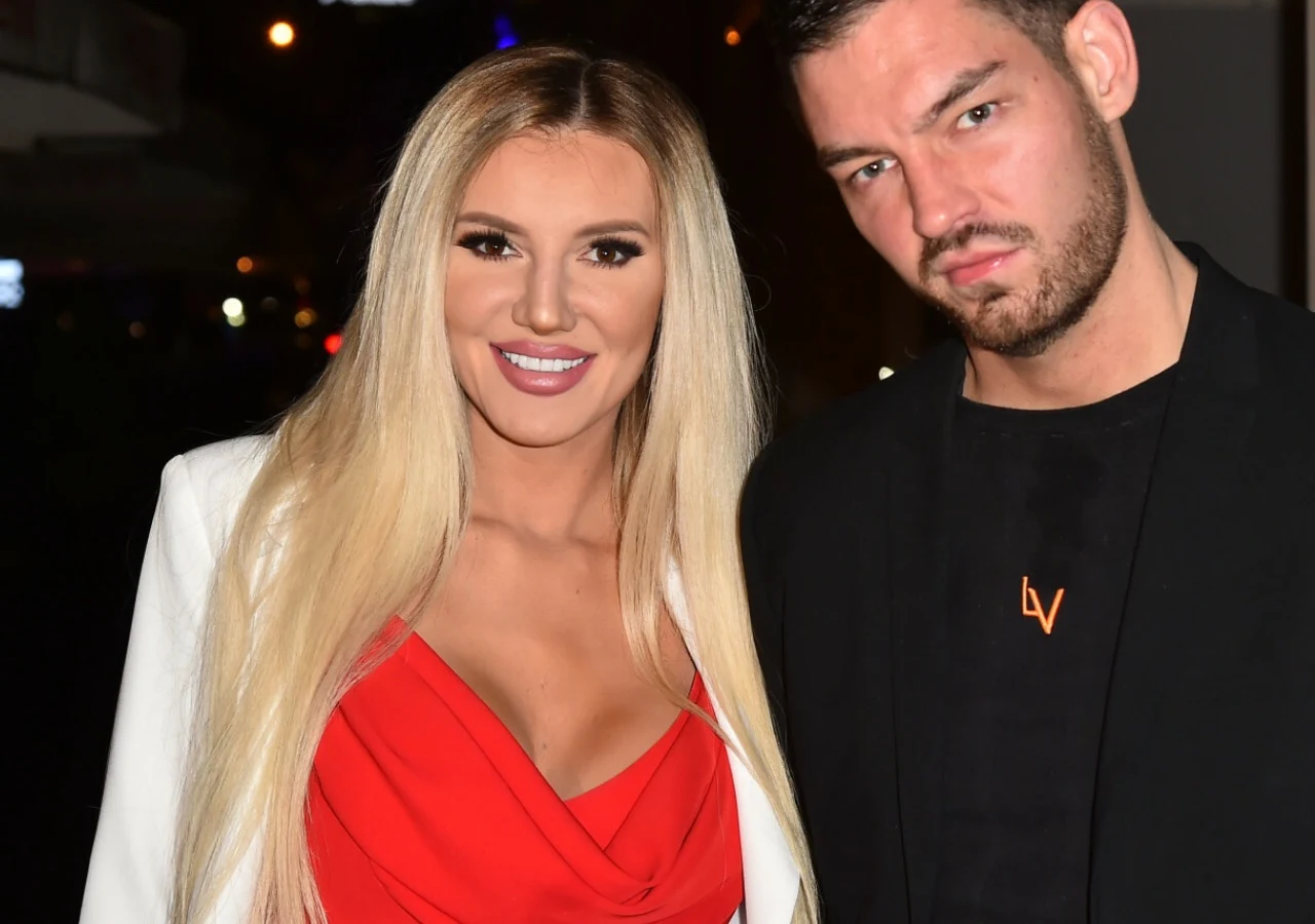 Kobieta z długimi blond włosami w czerwonej sukience i białej marynarce stoi obok mężczyzny w czarnej marynarce, oboje zwróceni w stronę aparatu.