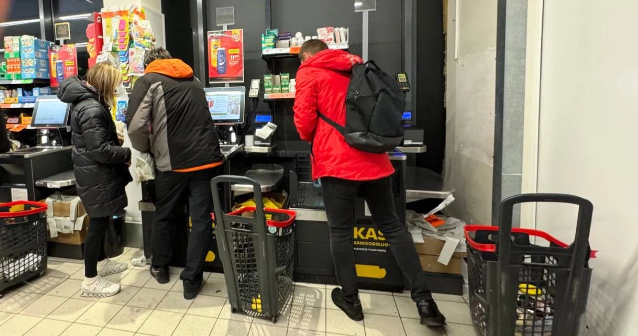 Biedronka do 18 lipca. Limit nawet 12 szt. Gratisy i największe promocje weekendu Trzy osoby w kurtkach przy samoobsługowych kasach w sklepie spożywczym z plastikowymi koszykami na zakupy.