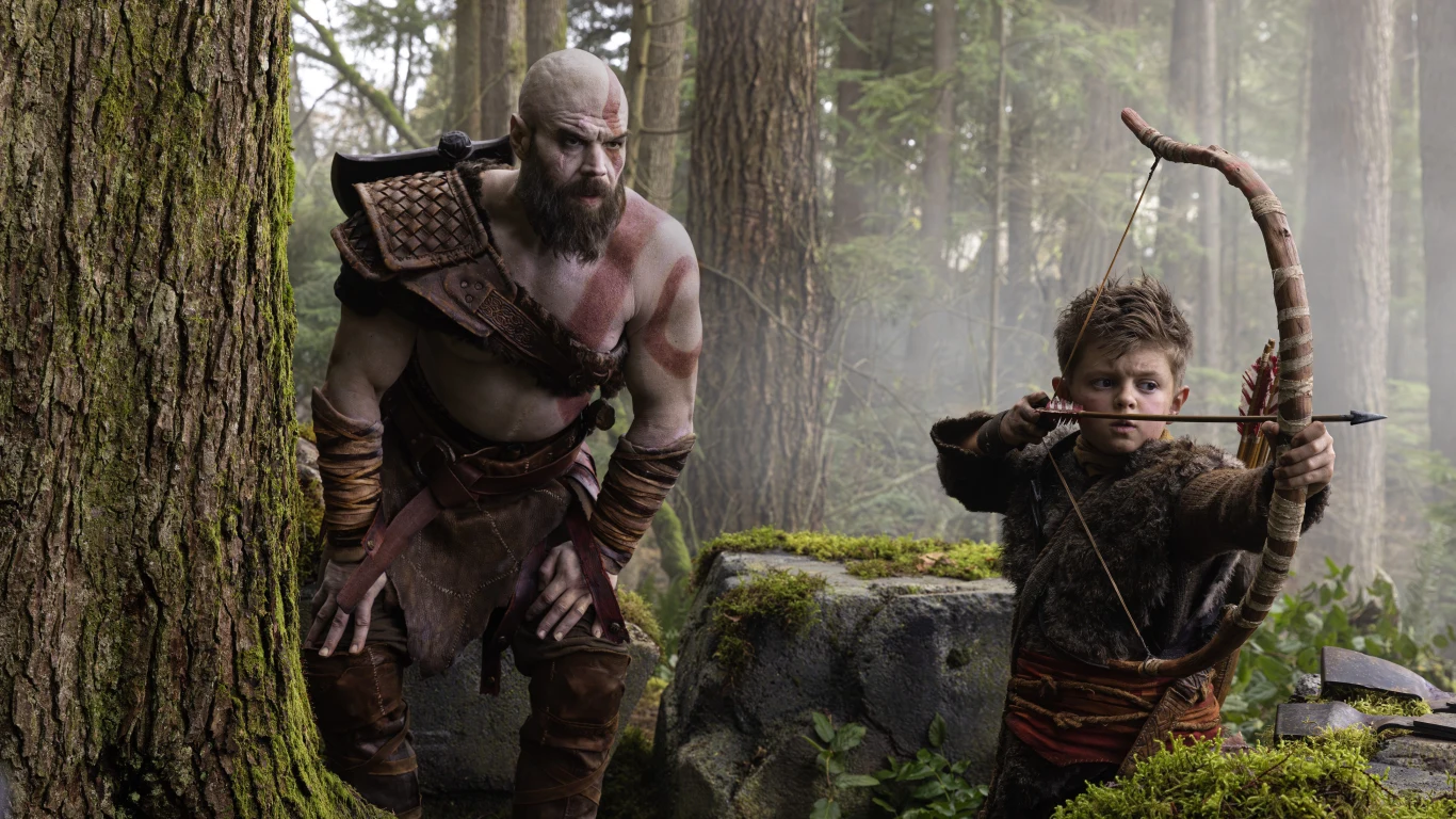 God of War - Ryan Hurst na pierwszym udostępnionym zdjęciu z serialu Ryan Hurst jako Kratos w skórzanej zbroi w lesie obok chłopca w futrzanej kamizelce z łukiem