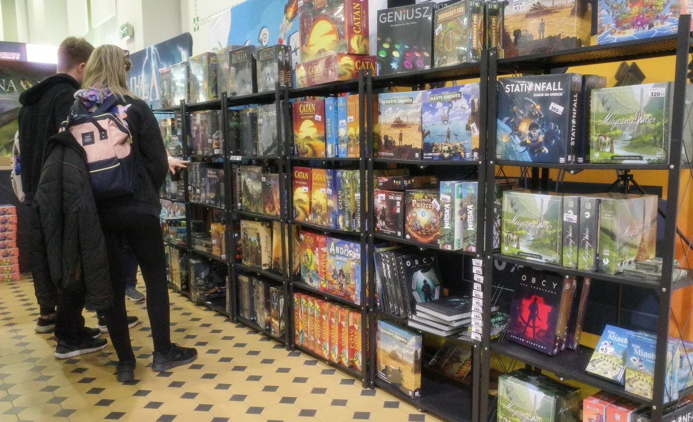Kolorowe pudełka z grami planszowymi ułożone na czarnych metalowych regałach, obok których stoją osoby przeglądające dostępne tytuły.
