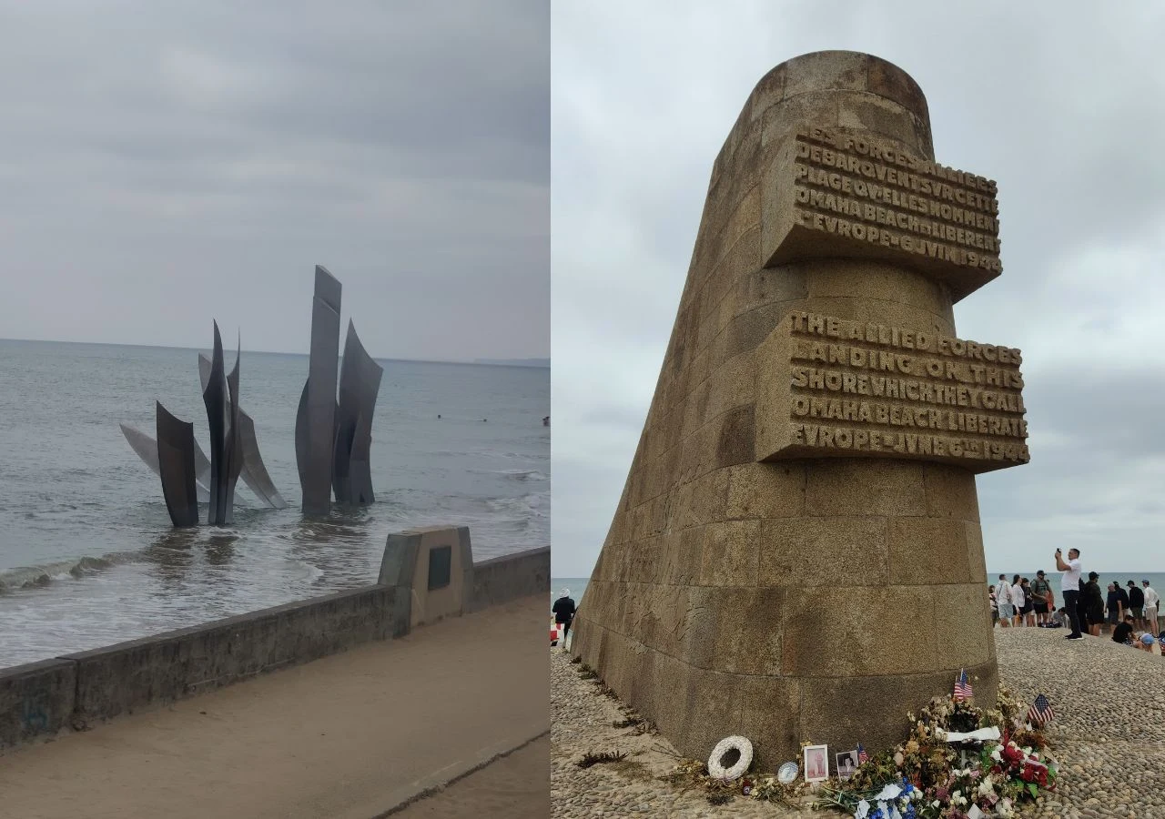 Pomnik upamiętniający lądowanie aliantów na plaży Omaha, nowoczesna instalacja stalowa wystająca z morza oraz monumentalna, kamienna rzeźba z wyrytym napisem, wokół której złożono kwiaty i symboliczną tabliczkę pamięci.