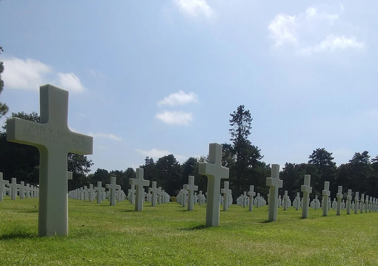 Rzędy białych krzyży ustawione na zielonej trawie cmentarza wojskowego, otoczone drzewami pod błękitnym niebem.