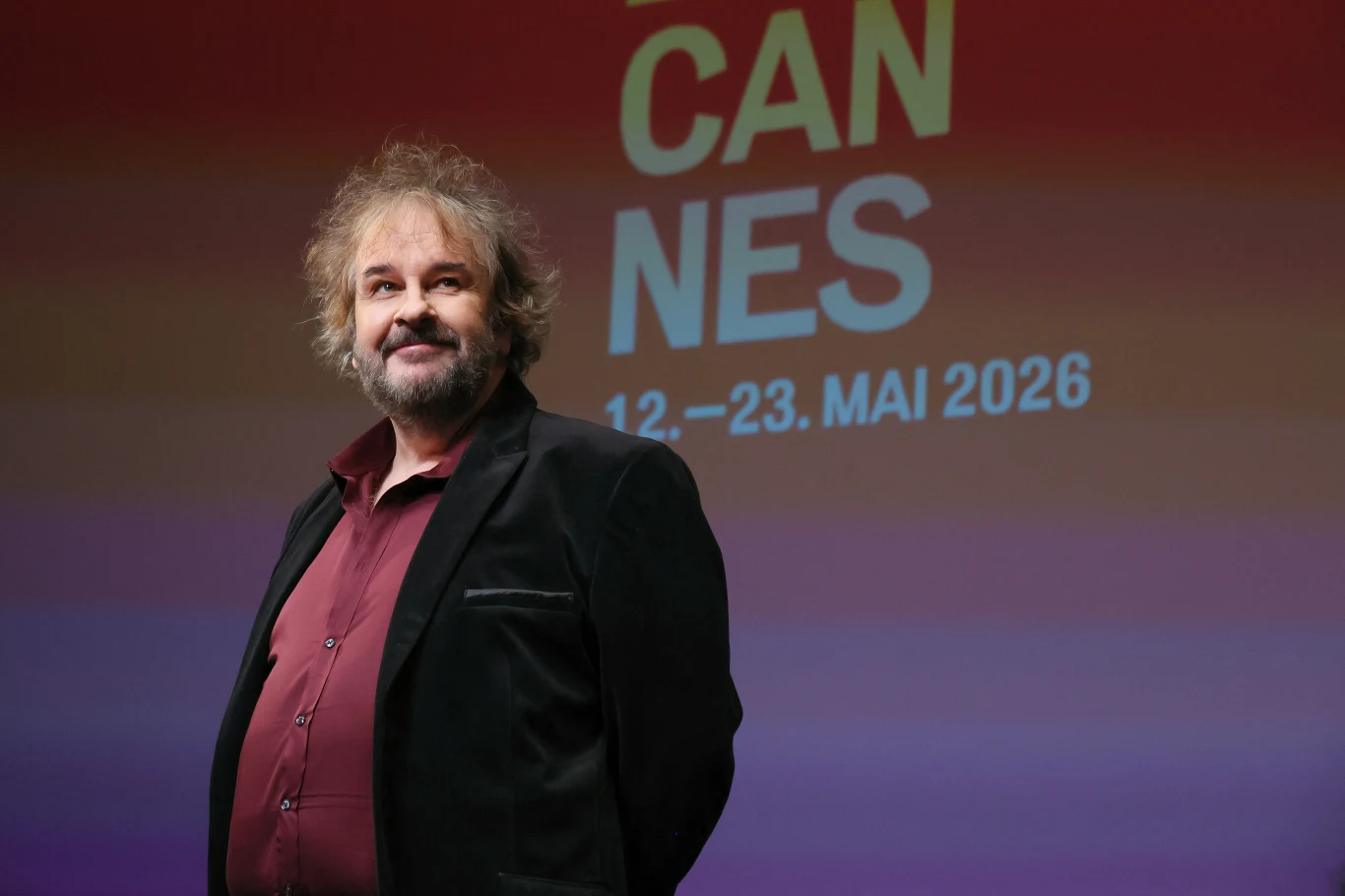 Peter Jackson Peter Jackson w ciemnej marynarce i bordowej koszuli na tle czerwonej planszy 'CANNES 12–23. MAI 2026'.